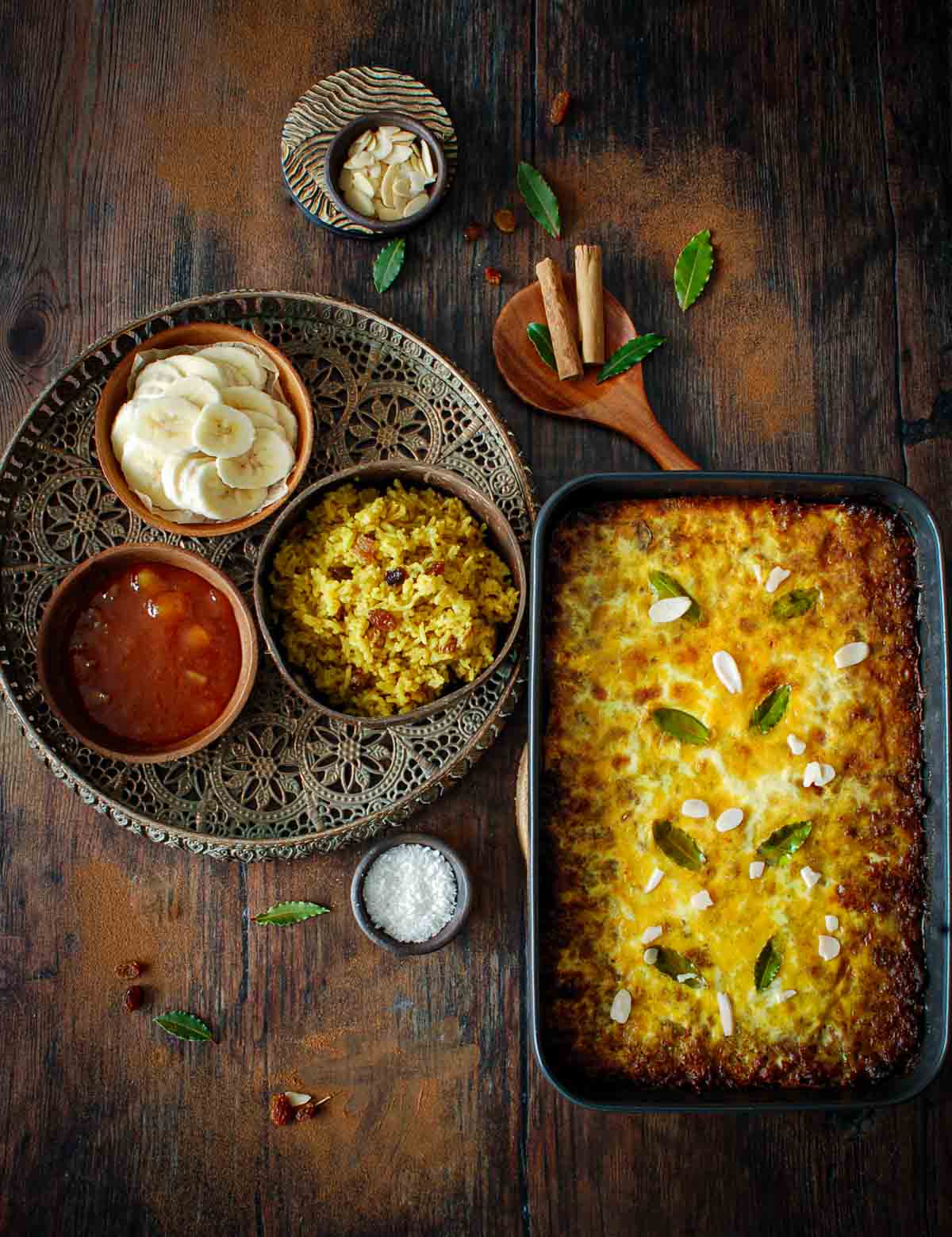 Cooked bobotie in an oven dish with side dishes of yellow rice, coconut, sliced banana and chutney