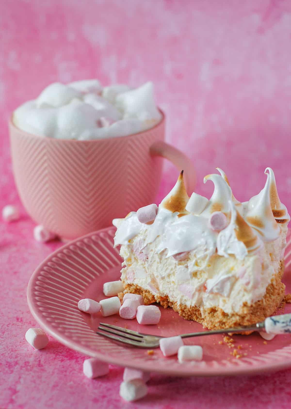 A slice of marshmallow tart on a pink plate with a mug of coffee in the background