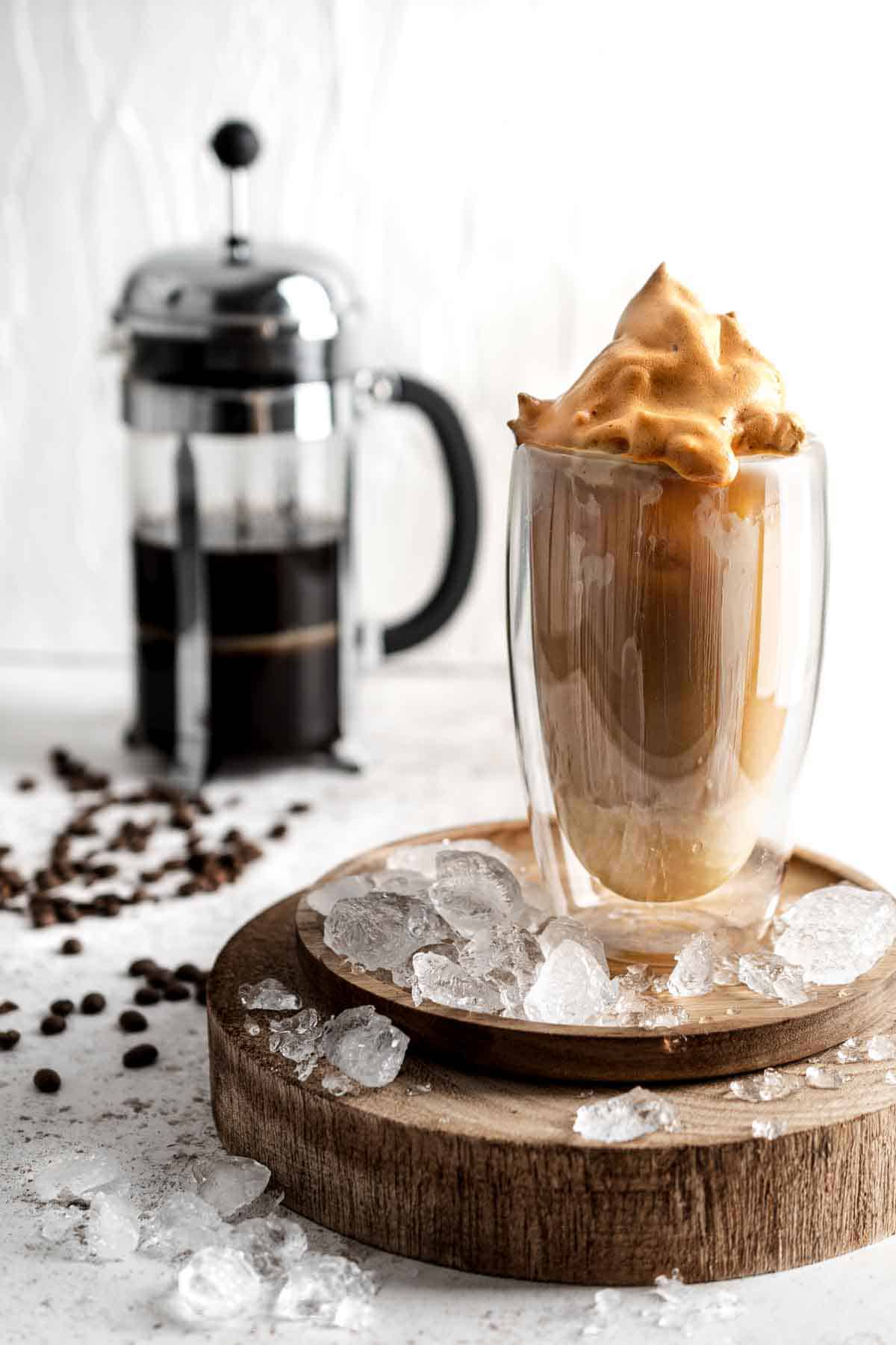 Tall glass of iced coffee on a wooden board, topped with Dalgona foam