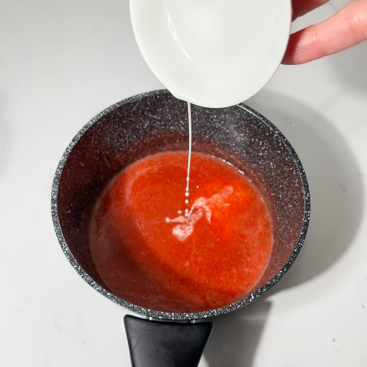 Adding cornstarch to the strawberry sauce in a small black saucepan.