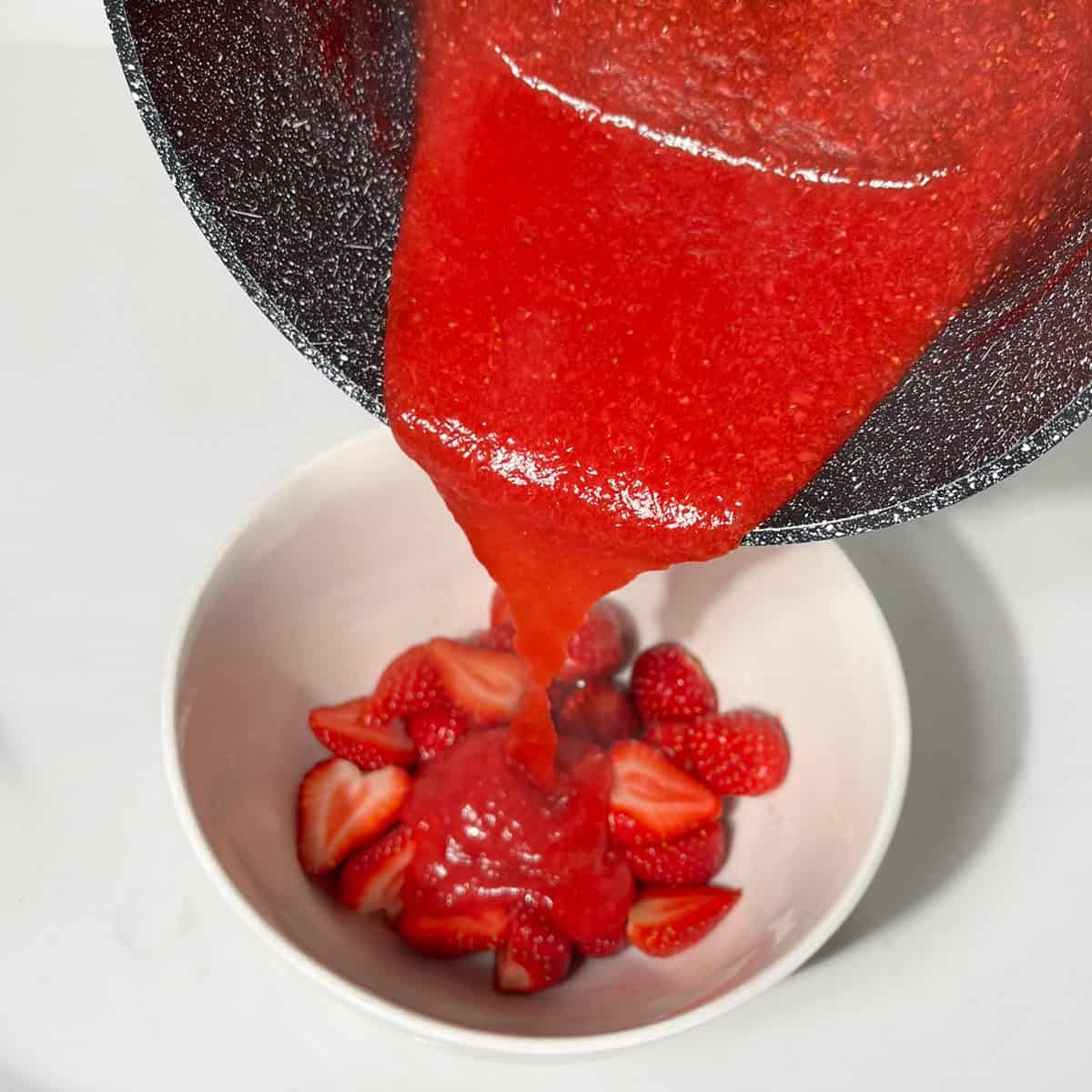 Pouring over the hot strawberry sauce over fresh, sliced strawberries in a white bowl.