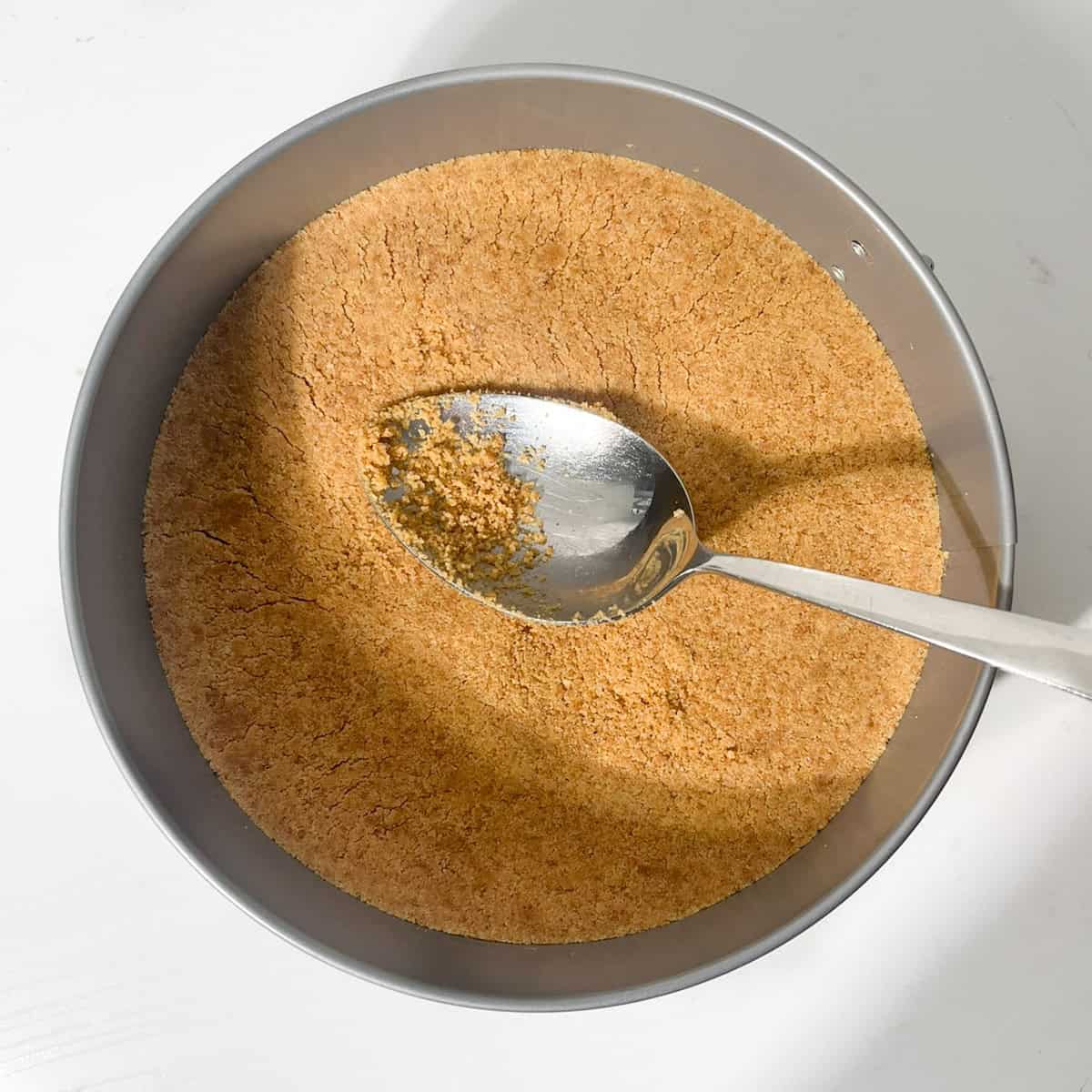Pressing the cookie crumbs and butter into the base of a springform tin with a large spoon.