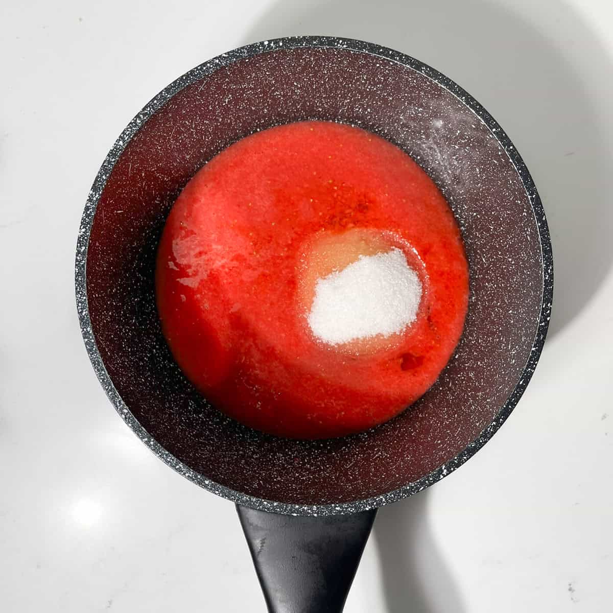 Adding sugar to the pureed strawberries in a small black saucepan.