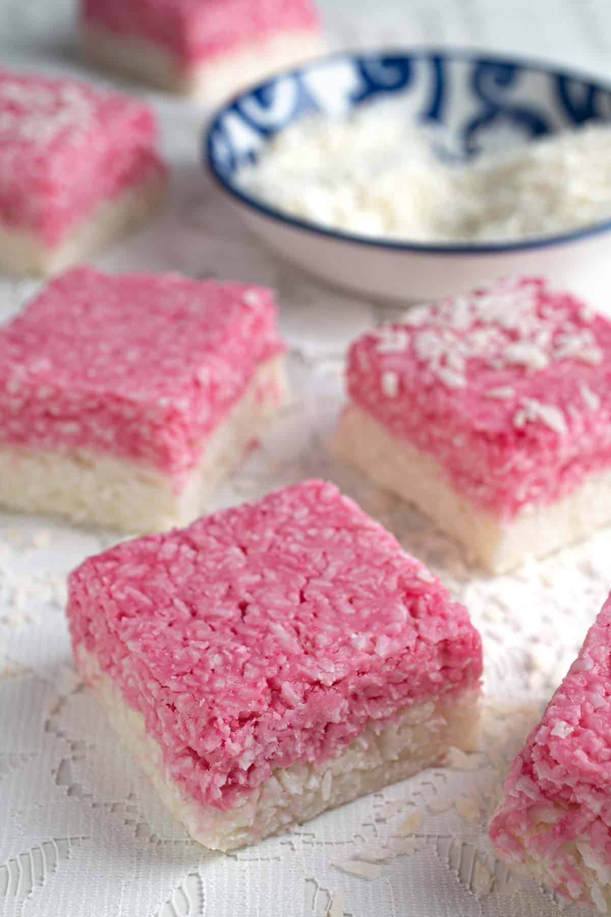 Pieces of white and pink coconut ice spread out on a table with a blue and white bowl in the background containing desiccated coconut. 