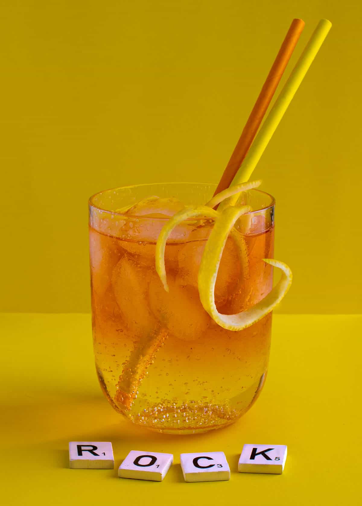 A medium glass of Rock shandy on a yellow background with an orange and yellow straw.