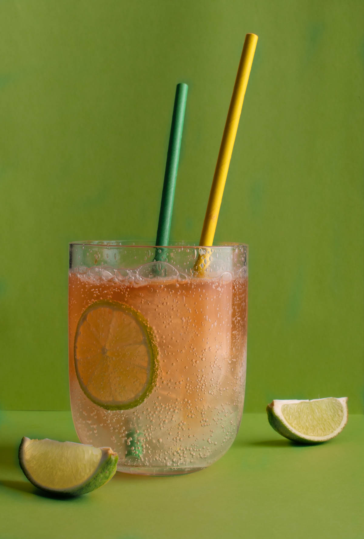 A glass of lemon, lime and bitters with two straws, on a green background.