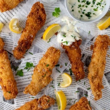 Fish goujons spread out on newspaper with some lemon slices and a garlic mayo dipping sauce on a small saucer.