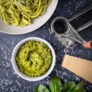 A bowl of pistachio peso surrounded by a bowl of pasta, a parmesan grater and parmesan cheese.