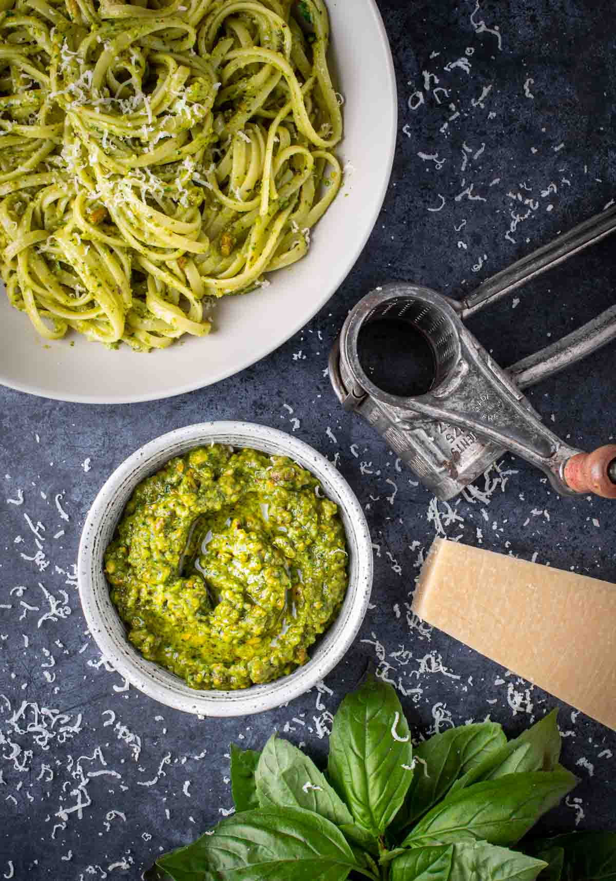 A bowl of pistachio peso surrounded by a bowl of pasta, a parmesan grater and parmesan cheese.