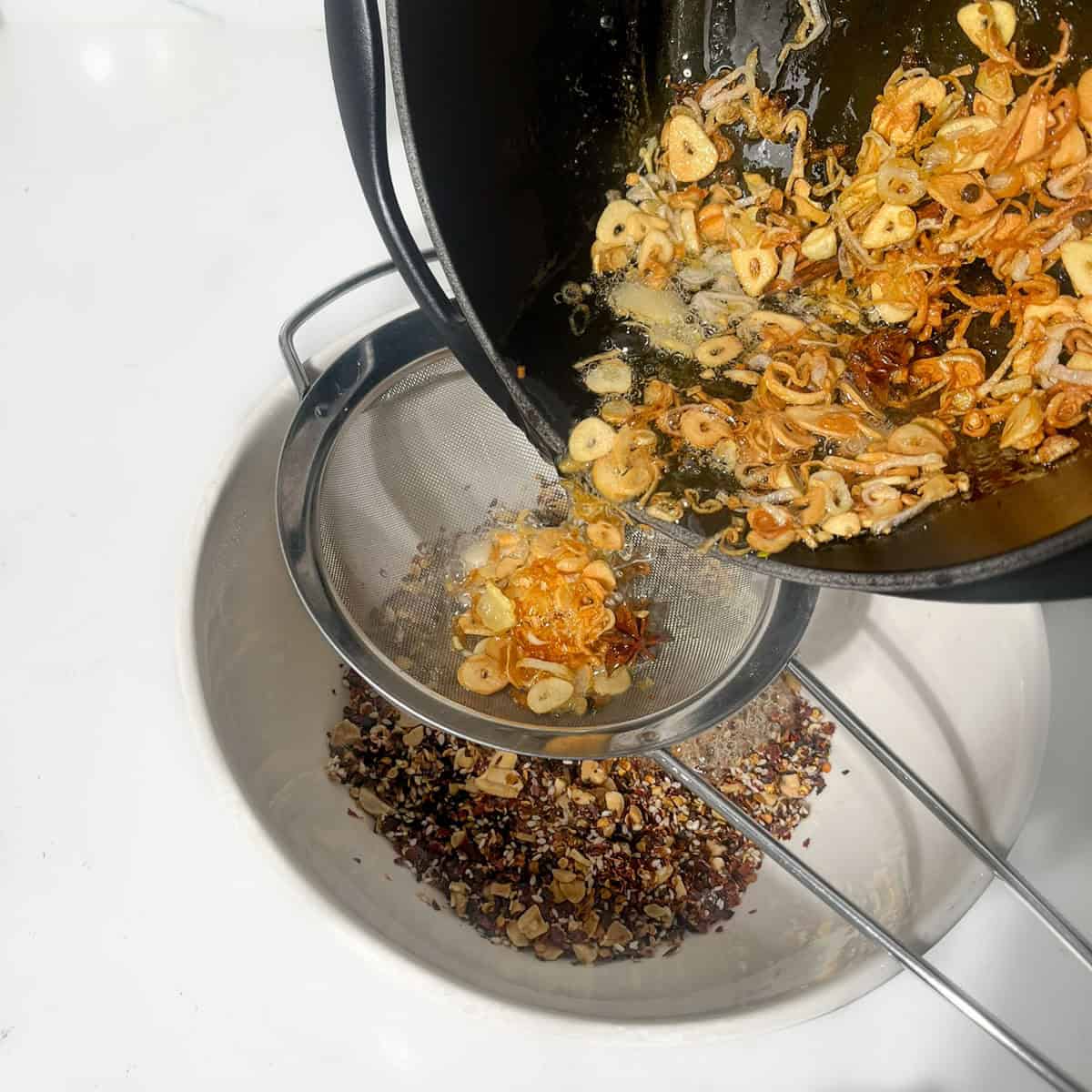 Pouring the oil through a sieve onto the dry chilli crisp oil ingredients in a large bowl.
