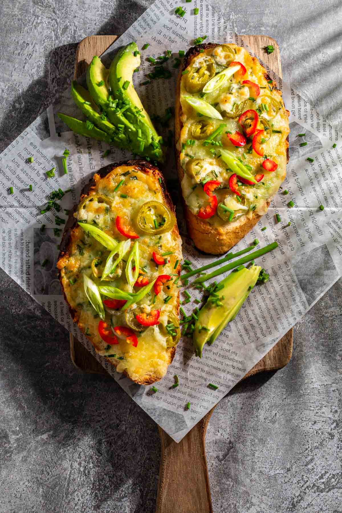 Two slices of cheese chilli toast on newspaper with a few slices of avocado.