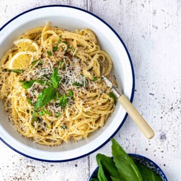 Lemon garlic pasta in a white enamel bowl, surrounded by a bowl of basil and a bowl of lemon slices.