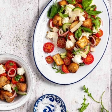 Panzanella toscana salad on a large white enamel serving dish, surrounded by a bowl of salad, salt and rocket leaves.