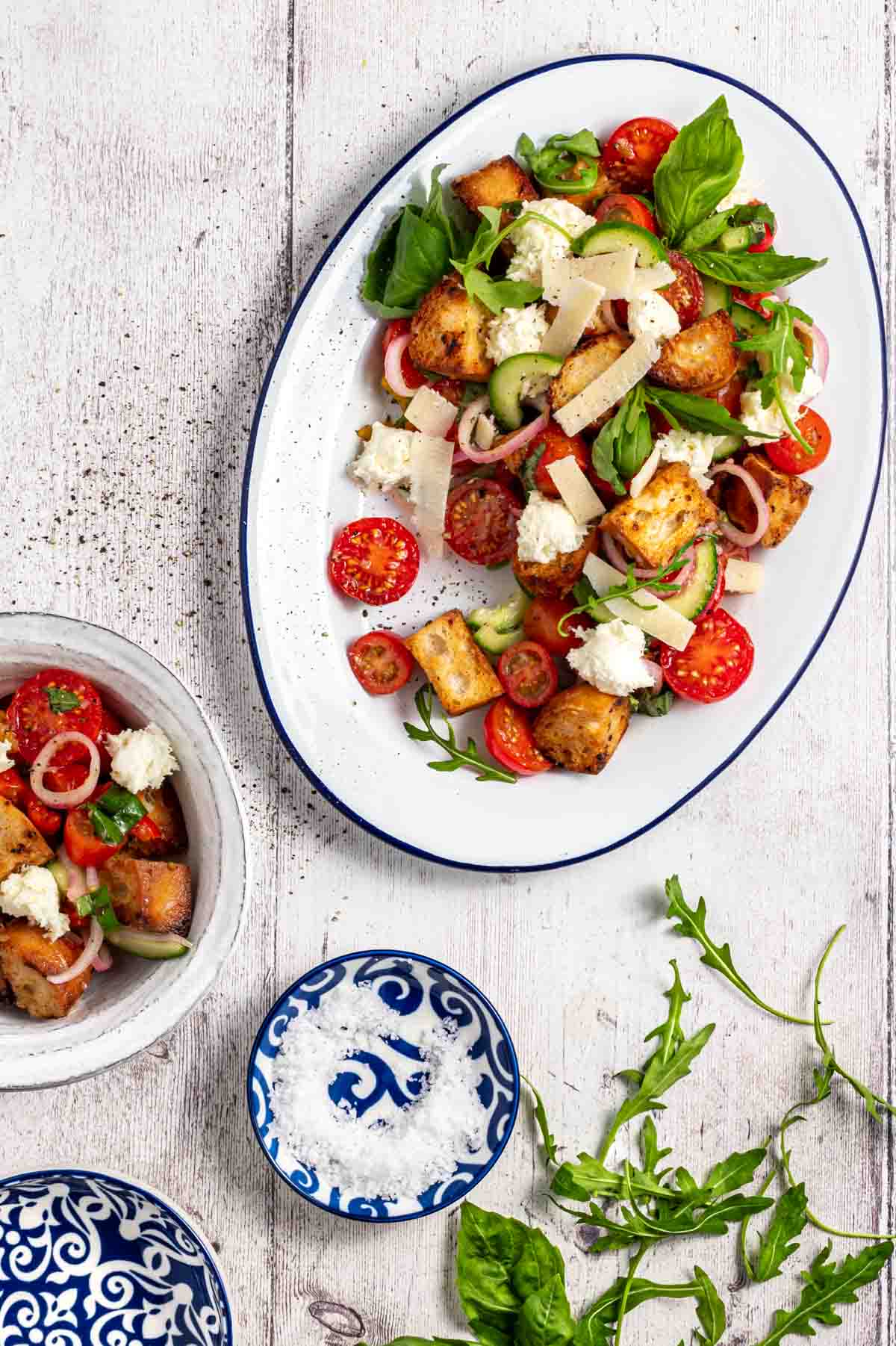 Panzanella toscana salad on a large white enamel serving dish, surrounded by a bowl of salad, salt and rocket leaves.