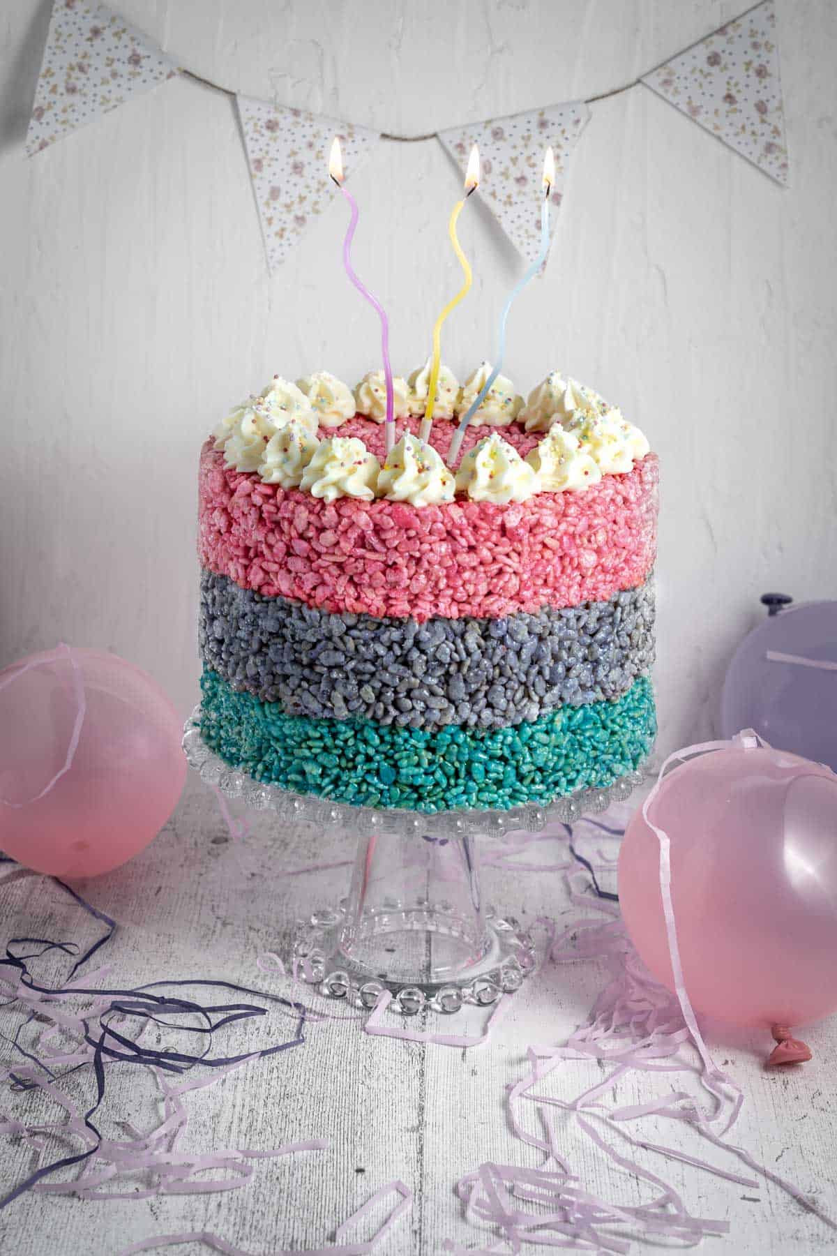 A three layered Rice Krispie cake on a cake stand, surrounded by balloons.