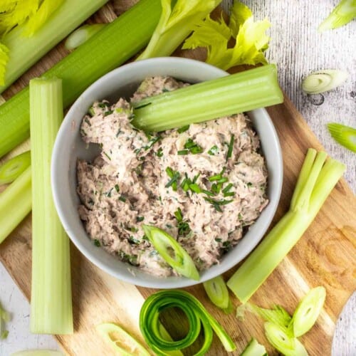 Tuna pate in a white bowl on a wooden serving board, surrounded by celery sticks.