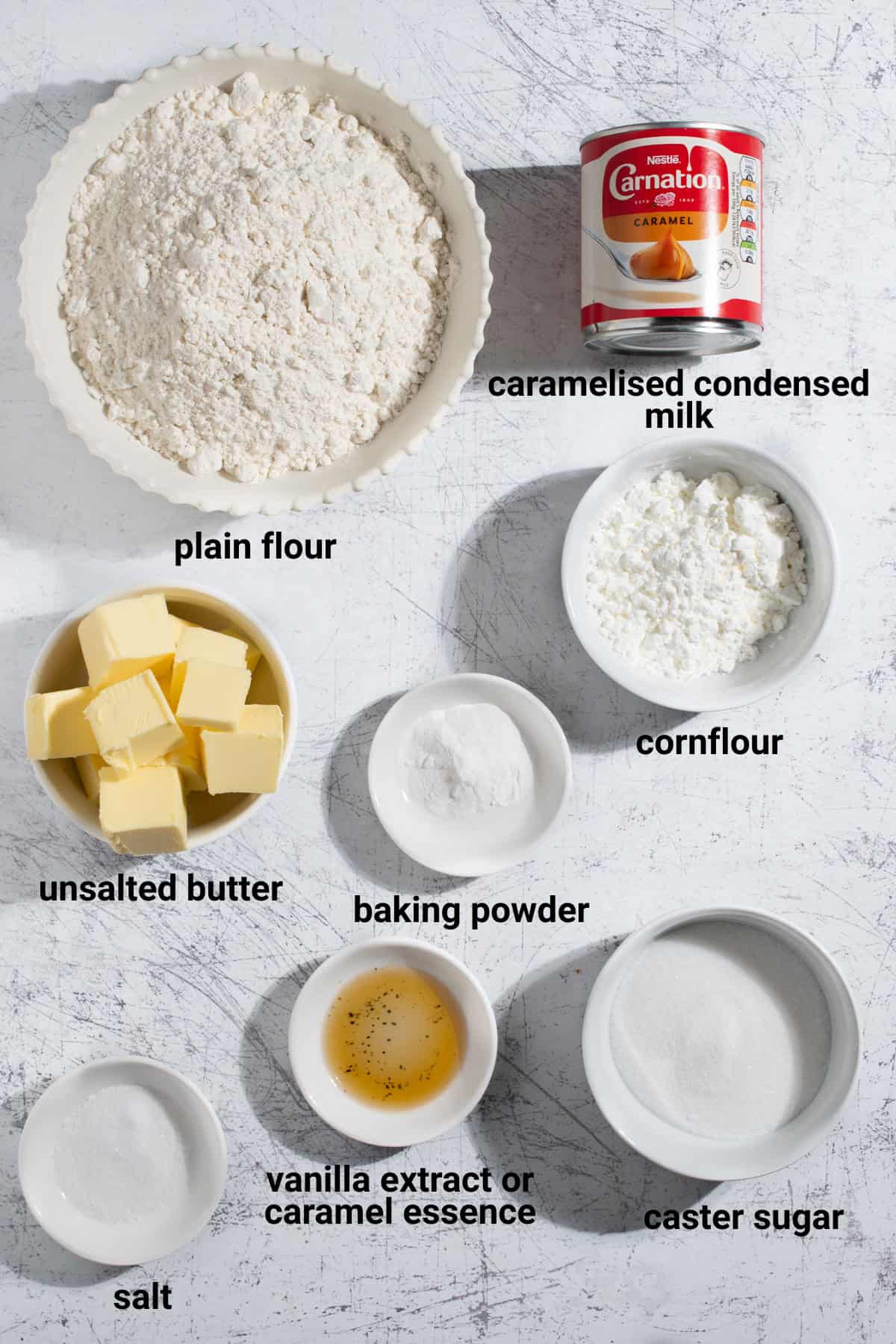 Ingredients for caramel condensed milk cookies arranged and labelled: plain flour, caramelised condensed milk, cornflour, butter, baking powder, vanilla or caramel essence, caster sugar and salt.