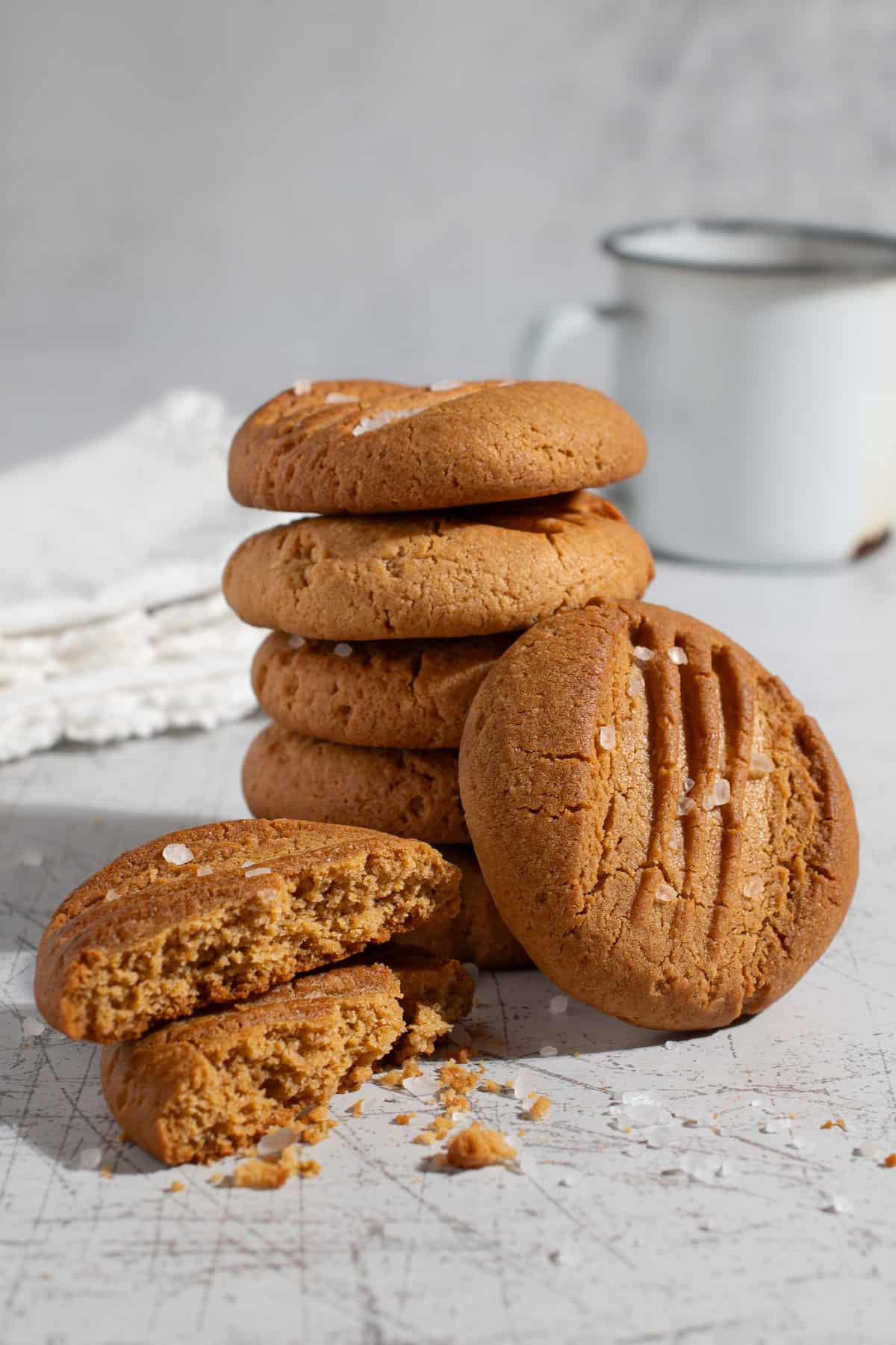 Stack of caramel condensed milk cookies with flaky sea salt, including one broken cookie showing the crumb and a white enamel mug in the background.