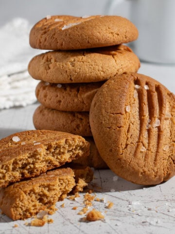 Stack of caramel condensed milk cookies with flaky sea salt, including one broken cookie showing the crumb and a white enamel mug in the background.