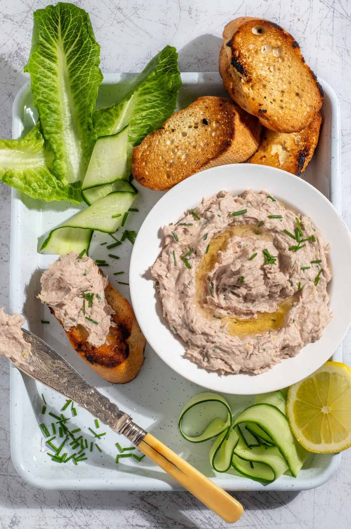 A bowl of tuna pate surrounded by toasted baguette, cucumber ribbons and lettuce.