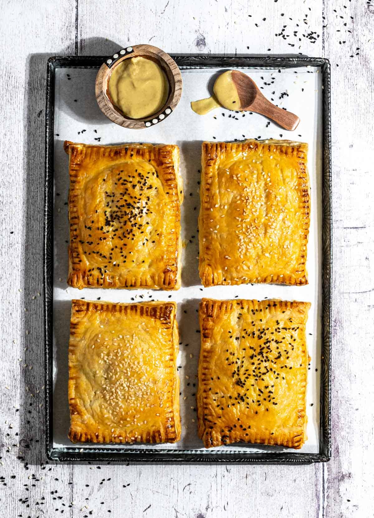 Four cheese and onion pasties on a baking sheet with a small bowl of mustard.