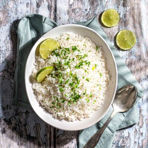 Coconut rice in a white bowl on a wooden background. The rice is topped with chopped coriander and slices of lime,