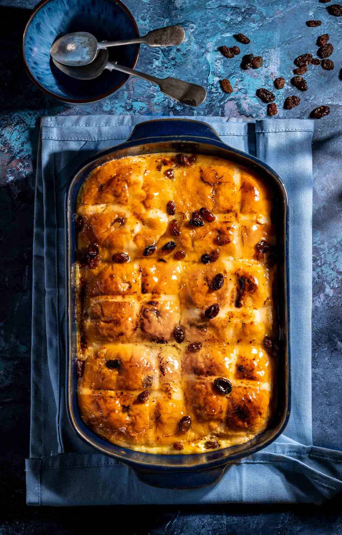 A hot cross bun bread and butter pudding in a blue baking dish, on a blue background.