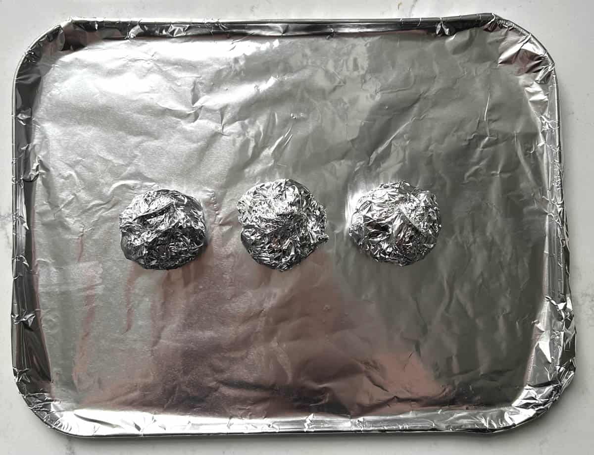 Three garlic heads wrapped in foil parcels on a baking sheet.