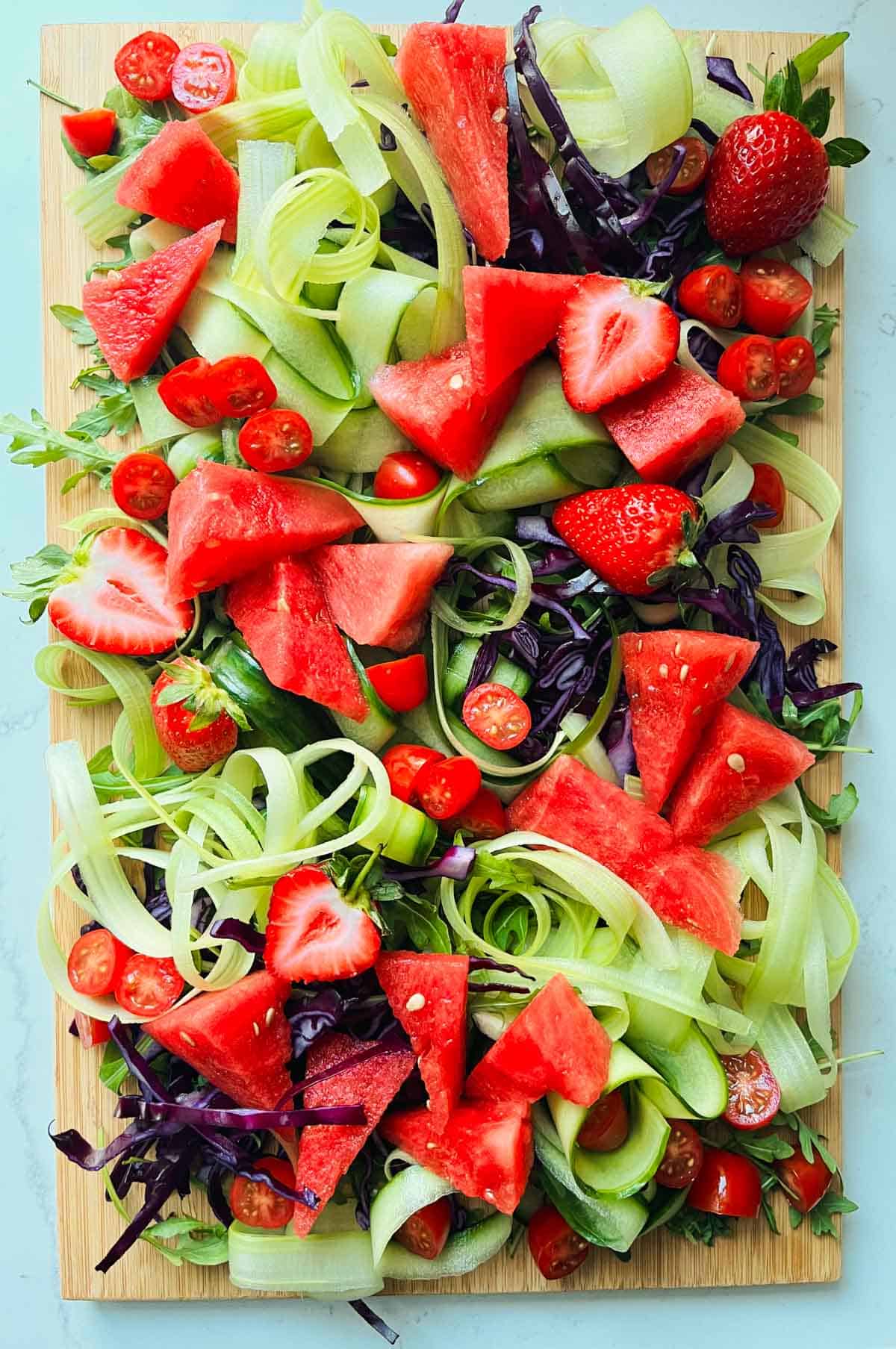 Rocket leaves, chopped red cabbage, celery and cucumber ribbons, watermelon, strawberries and tomatoes on a wooded board.