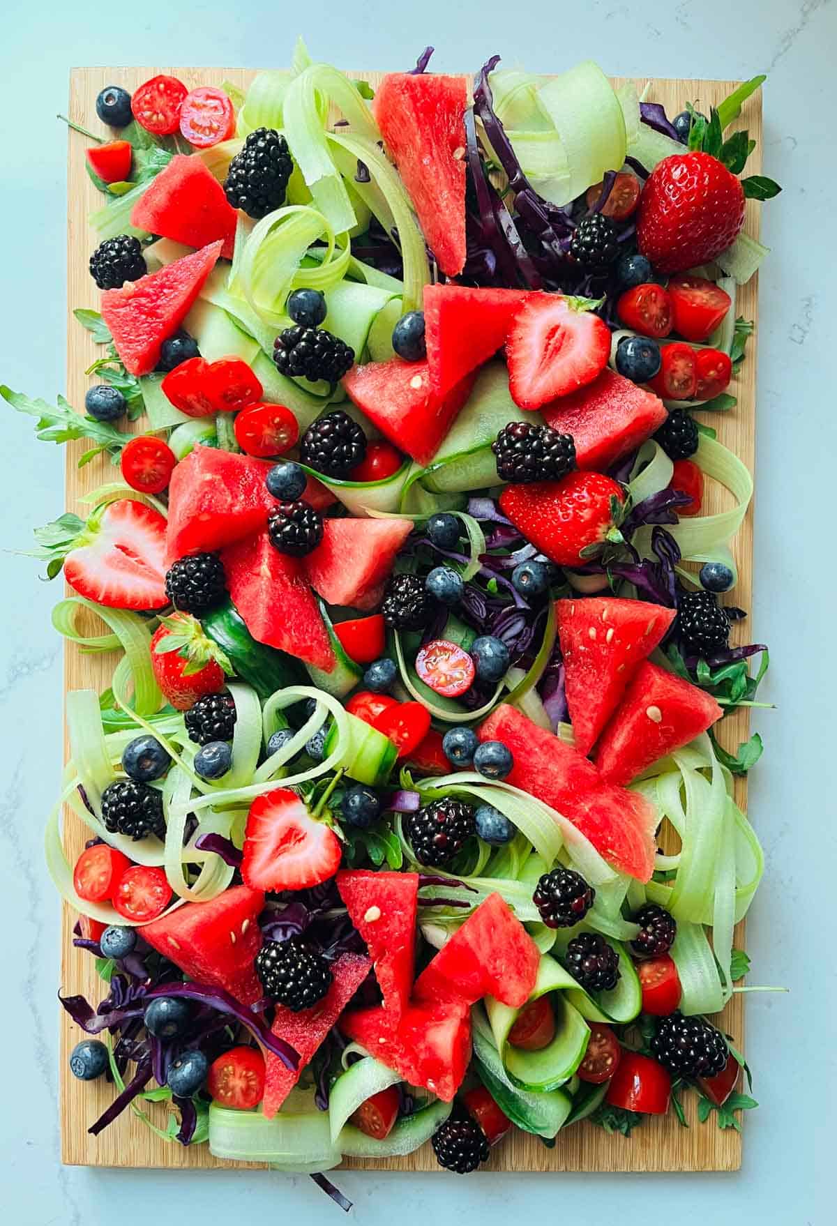 Rocket leaves, chopped red cabbage, celery and cucumber ribbons, watermelon, strawberries, tomatoes, blackberries and blueberries on a wooded board.