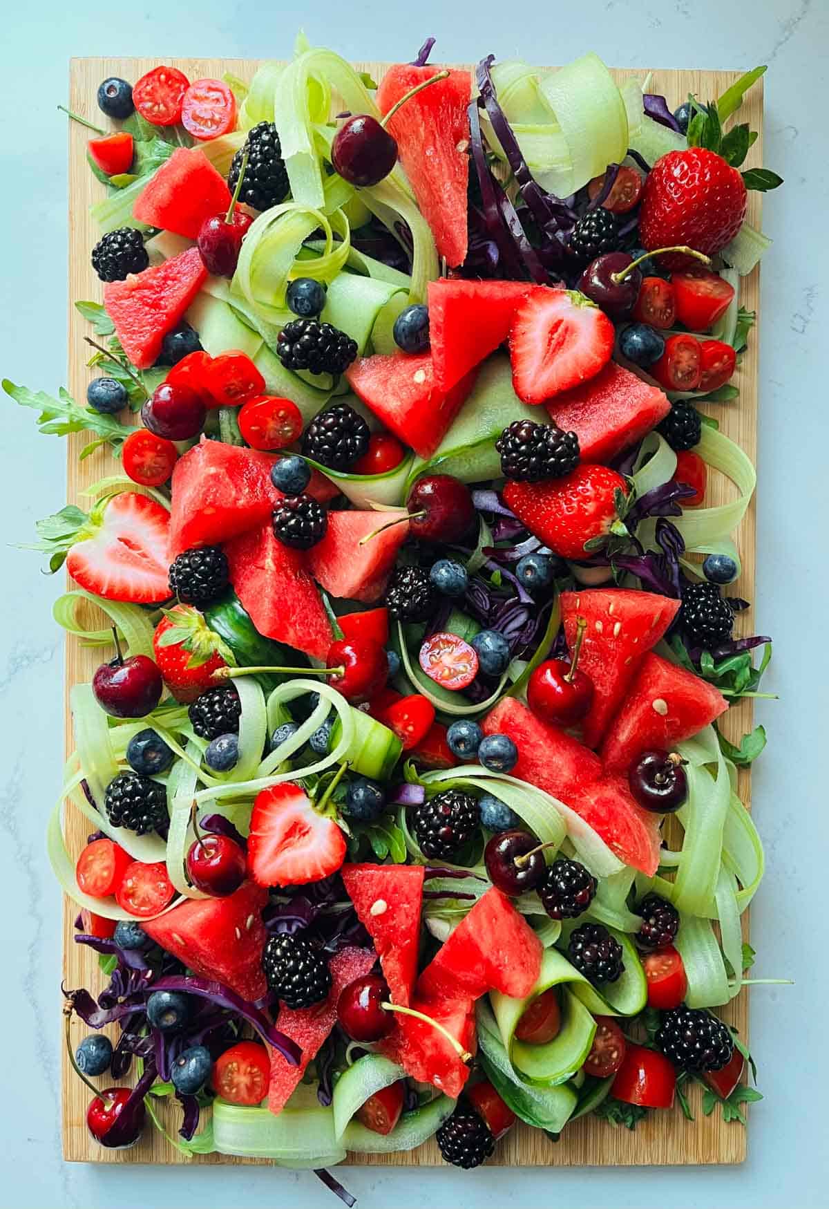 Rocket leaves, chopped red cabbage, celery and cucumber ribbons, watermelon, strawberries, tomatoes, blackberries, blueberries and cherries on a wooded board.
