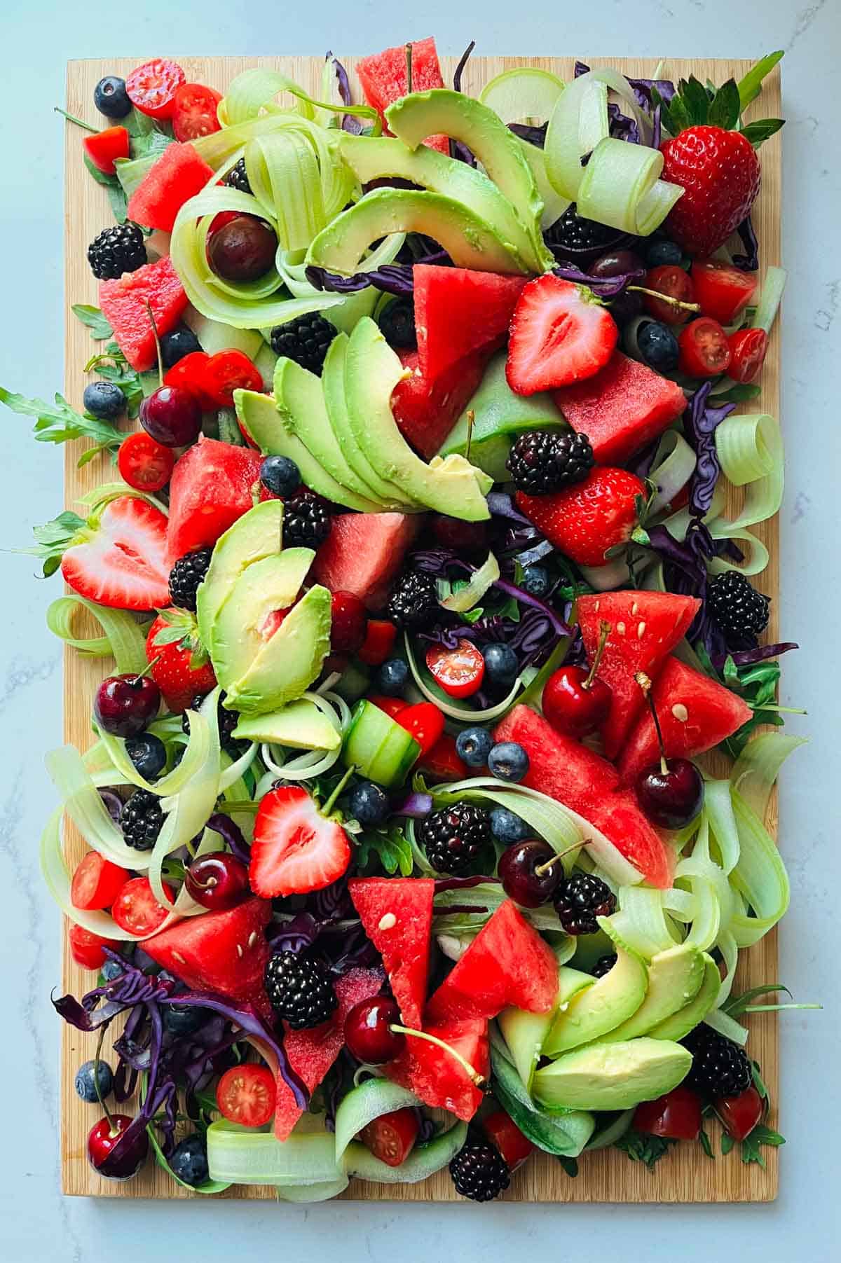 Rocket leaves, chopped red cabbage, celery and cucumber ribbons, watermelon, strawberries, tomatoes, blackberries, blueberries, cherries and avocado on a wooded board.