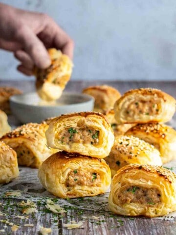 Chicken sausage rolls scattered on a table with a hand in the background dipping a sausage roll into sauce.