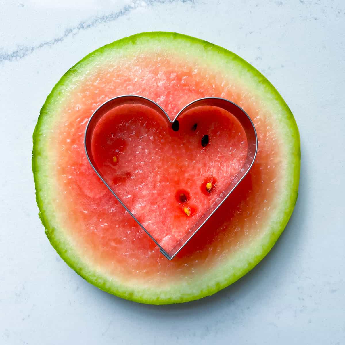 A heart shaped cookie cutter resting on top of a slice of watermelon.