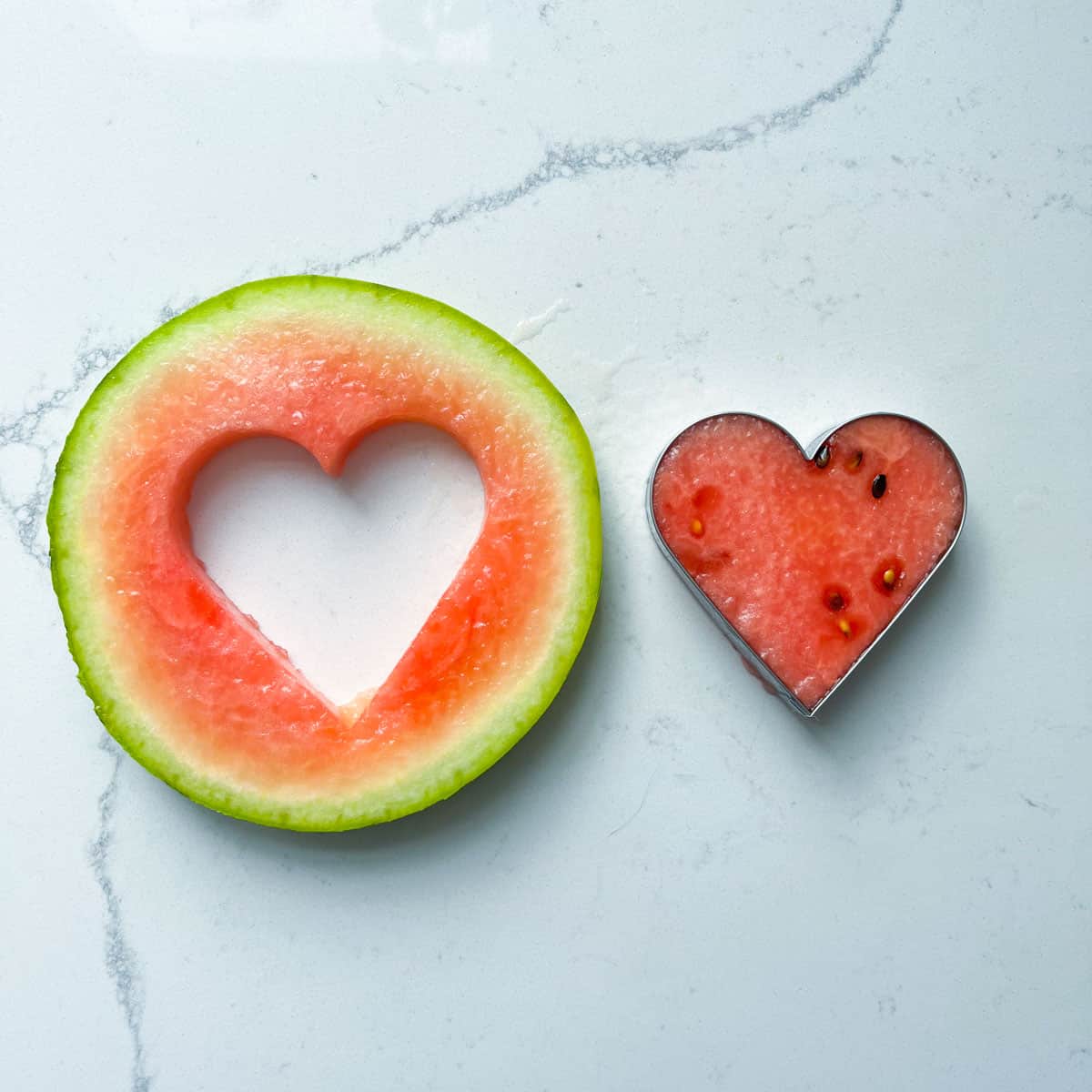 A heart shaped piece of watermelon cut from a larger piece of watermelon.