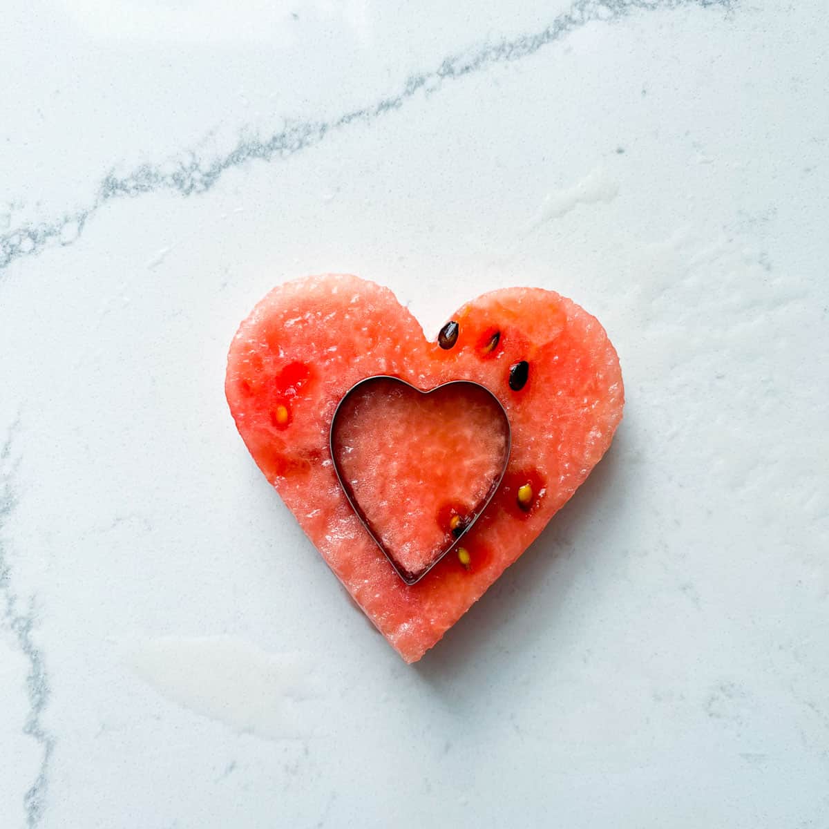 A small heart shaped cookie cutter resting on top of a larger heart shaped piece of watermelon.