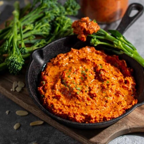 Romesco dip in a small black pan surrounded by broccoli spears, with a bottle of sauce in the background.