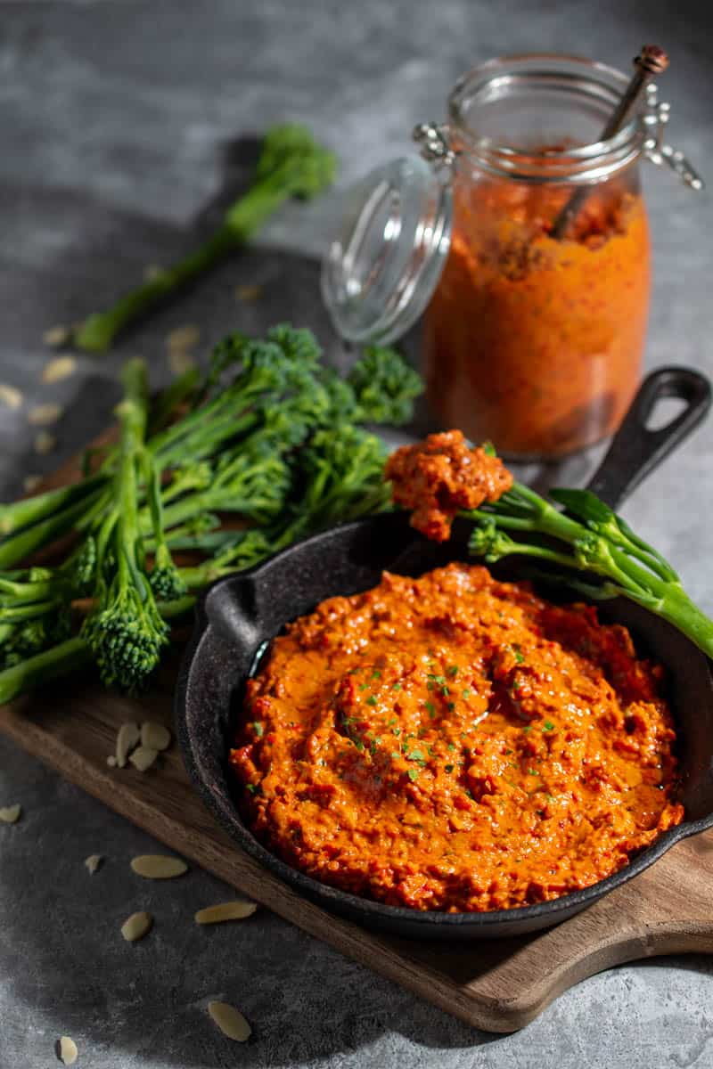 Romesco dip in a small black pan surrounded by broccoli spears, with a bottle of sauce in the background.