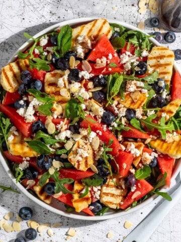 Grilled halloumi and watermelon salad on a large white salad plate with blueberries scattered on the surface around it.
