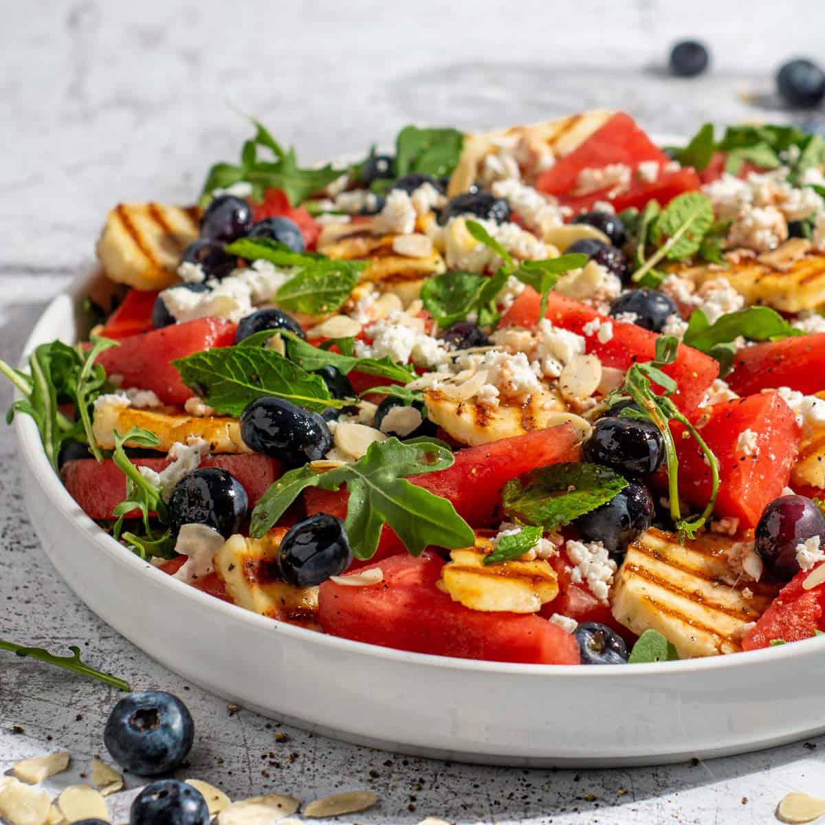 Grilled halloumi and watermelon salad on a large white salad plate with blueberries scattered on the surface around it.