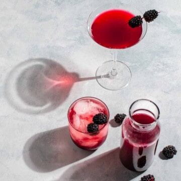 Blackberry simple syrup in a carafe next to a glass of blackberry syrup with lemonade and a blackberry martini.