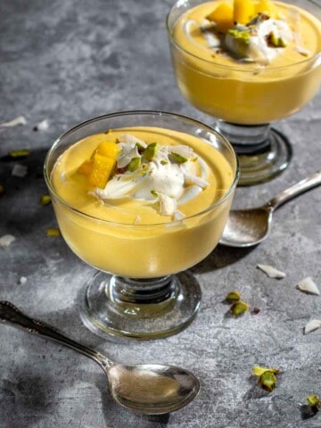Two parfait glasses with mango mousse, topped with cream and nuts on a grey background.