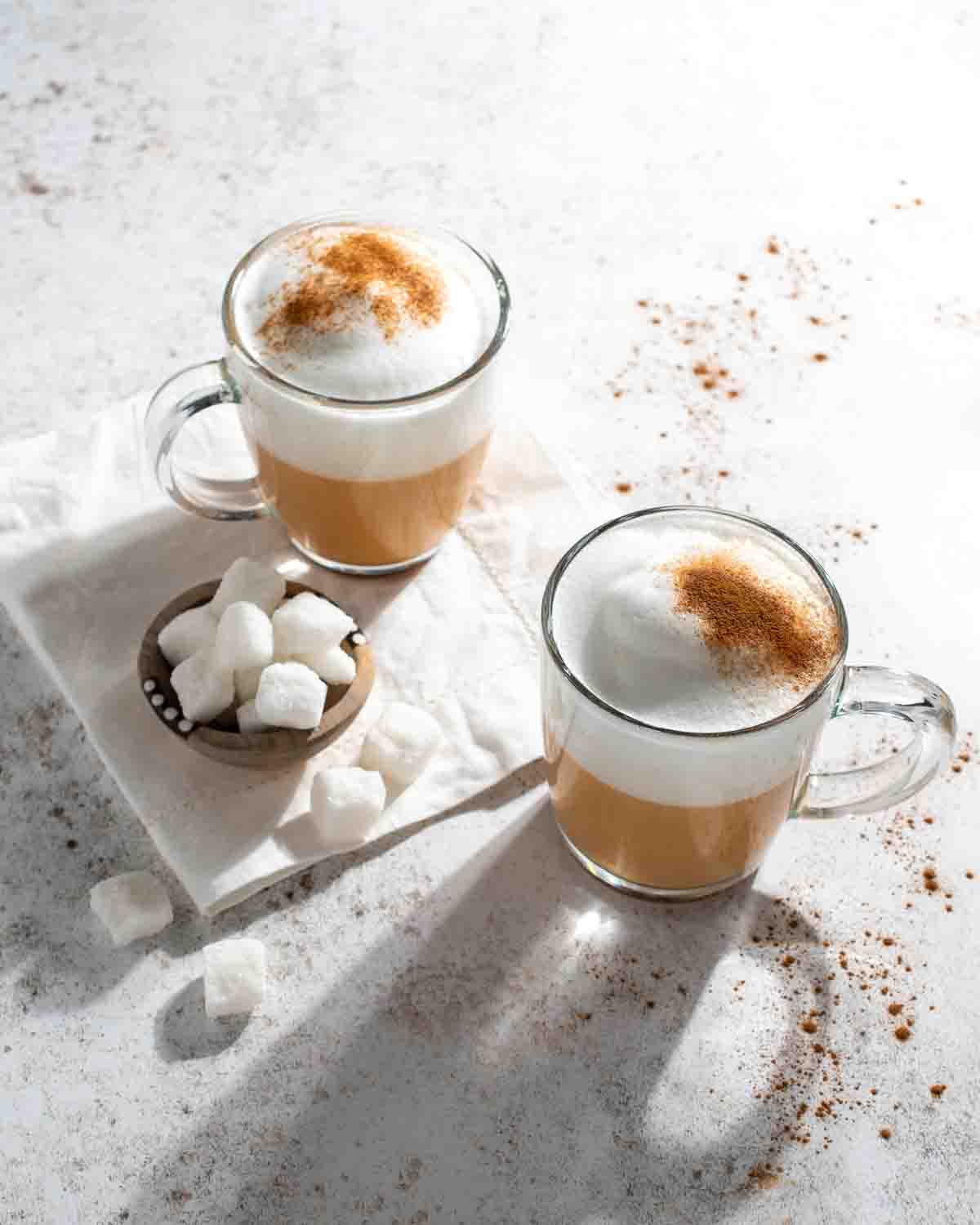 Two mugs of red cappuccino with some sugar cubes in a small bowl.