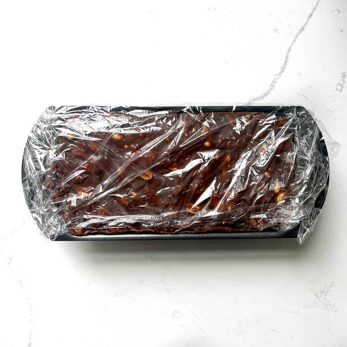 Tinginys mixture in a loaf pan covered with plastic wrap.