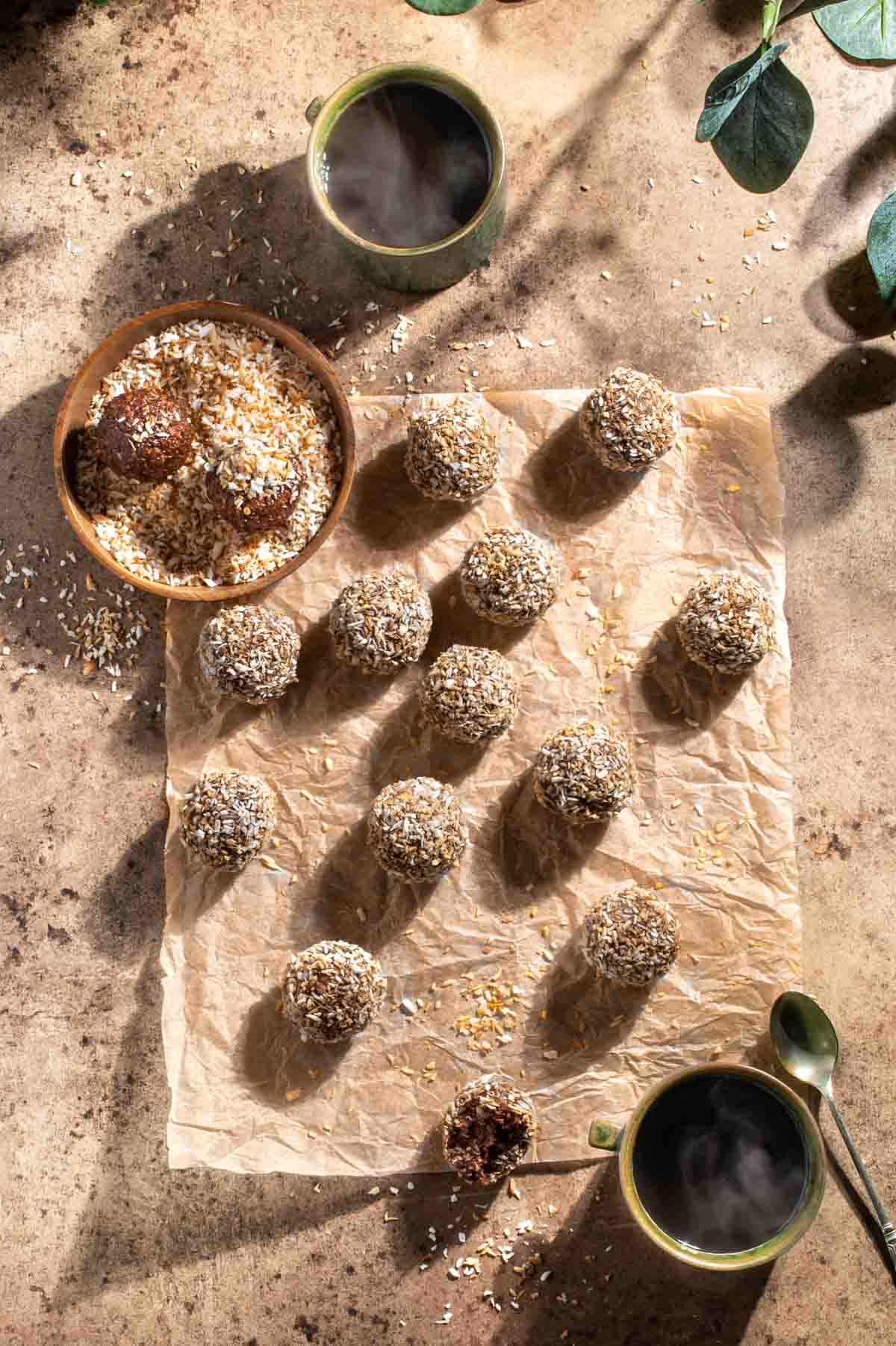 Chokladbollar on a piece of brown paper surrounded by a bowl of desiccated coconut and two mugs of coffee.