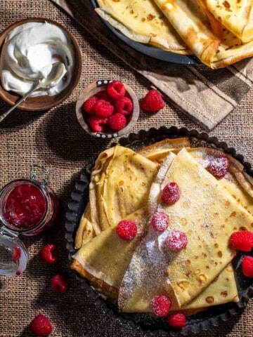 Pandekager folded in triangles in a black, round backing dish, served with raspberries, icing sugar, jam and cream.