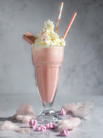 A large glass of strawberry milkshake with whipped cream in a milkshake glass surrounded by cotton candy.