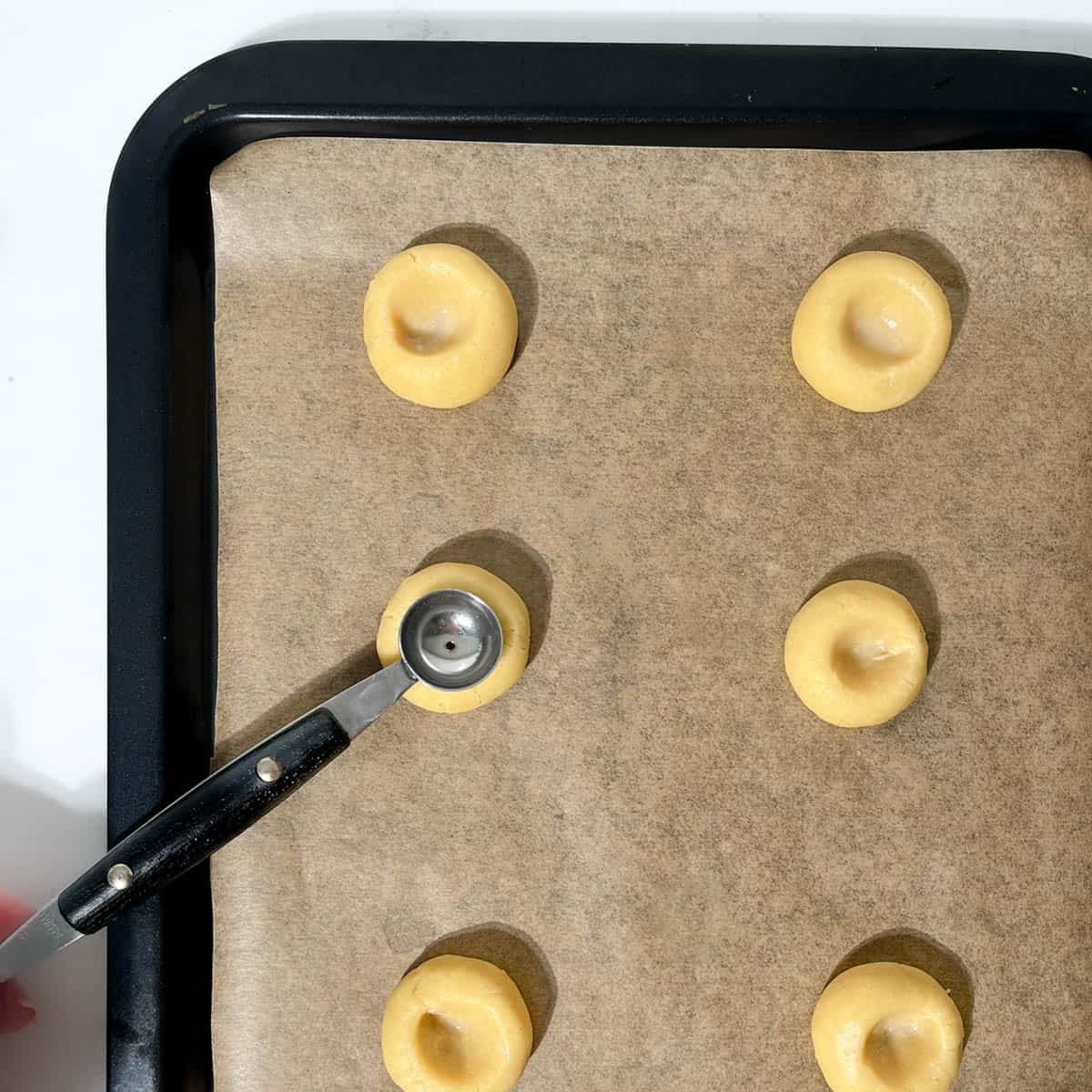 Indenting round dough balls for the thumbprint icing cookies with a small melon baller on a baking tray.