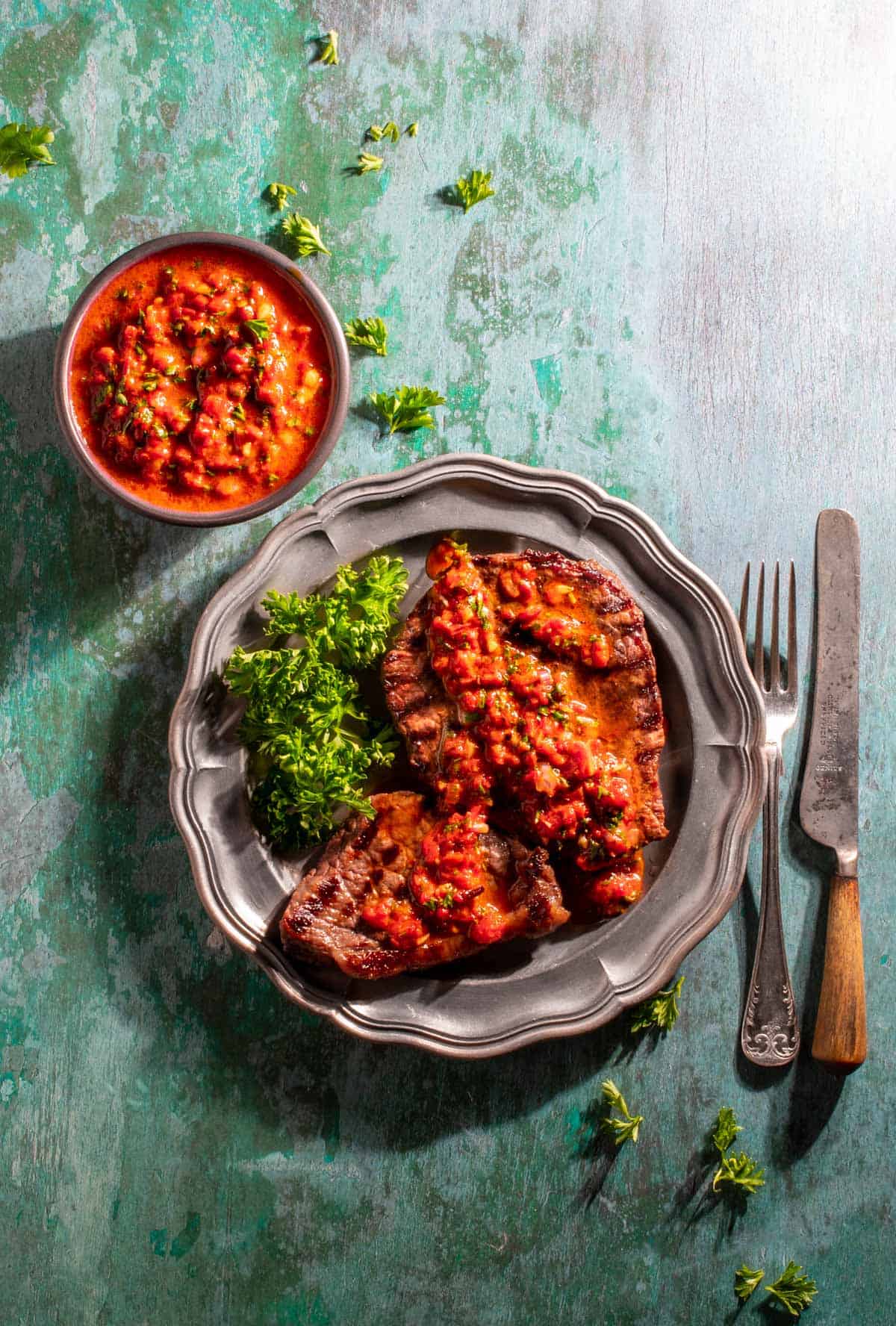 Red chimichurri in a bowl next to a plate with two steaks, with Red chimichurri spread onto it. 