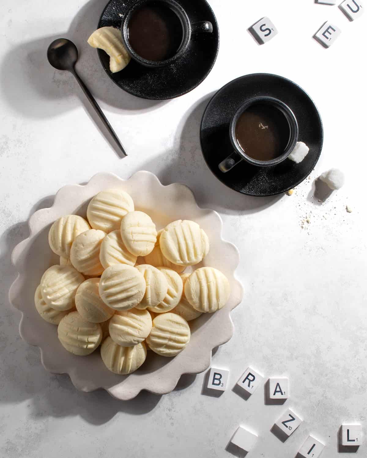 Sequilhos cookies on a white plate with two cups of coffee and some scrabble pieces. 